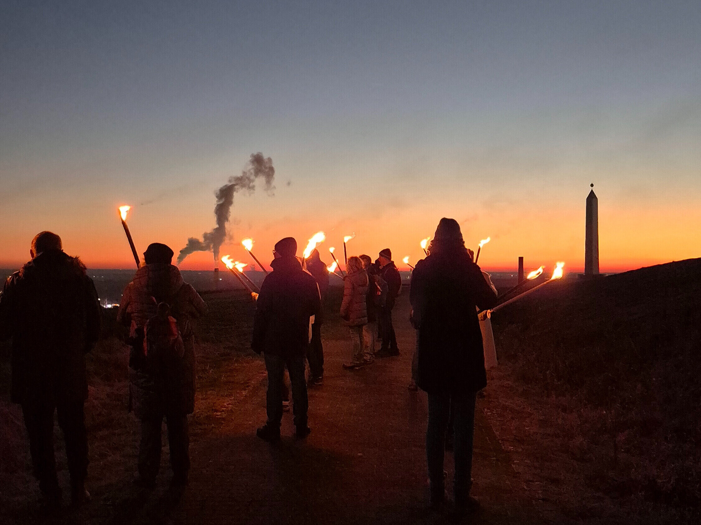 Eine Gruppe Menschen mit Fackeln läuft im Sonnenuntergang Richtung Sonnenuhr der Halde Hoheward.