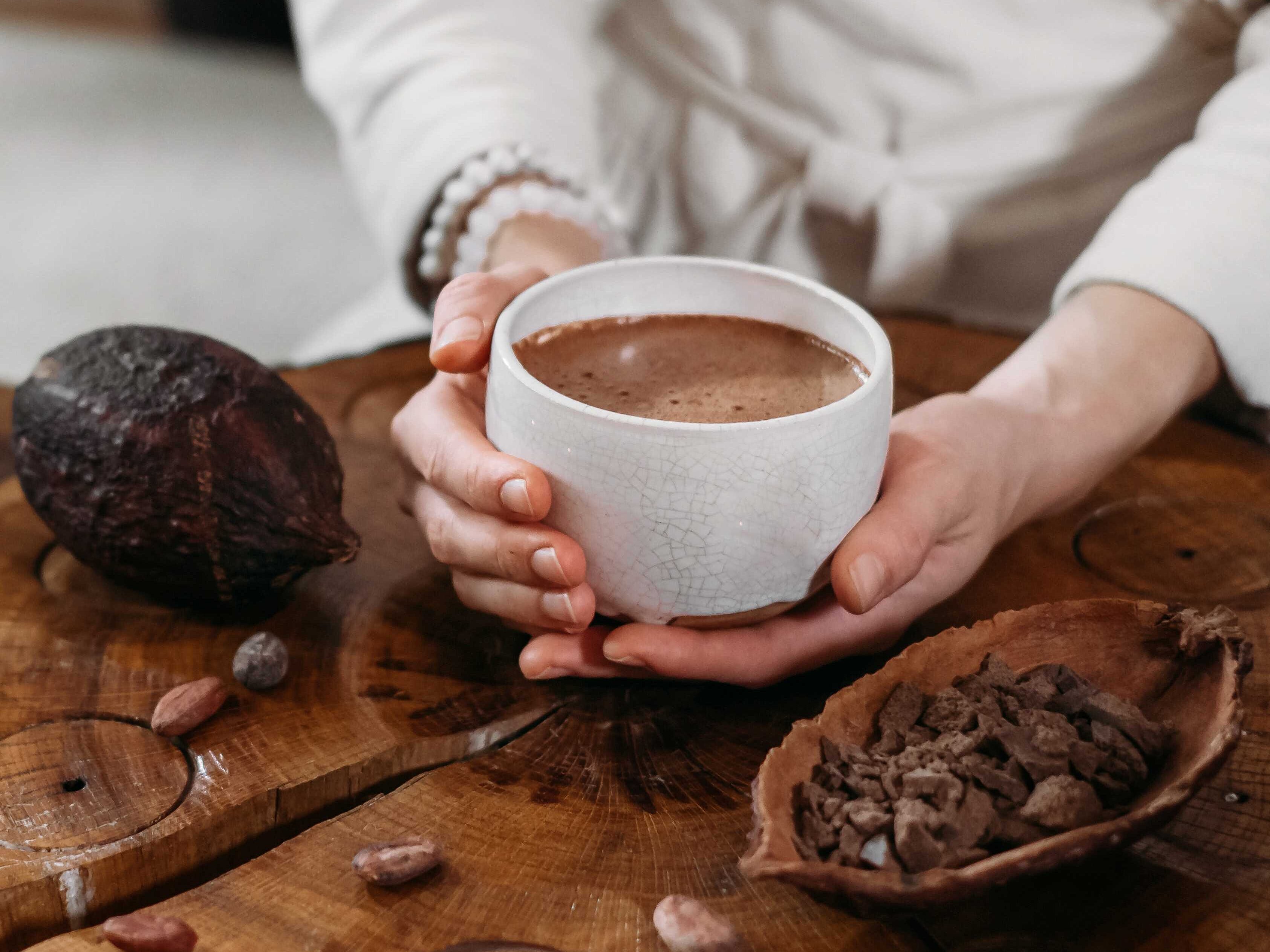 Eine Person hält eine Tasse mit Kakao in ihren Händen. Neben ihr liegen Kakeobohnen