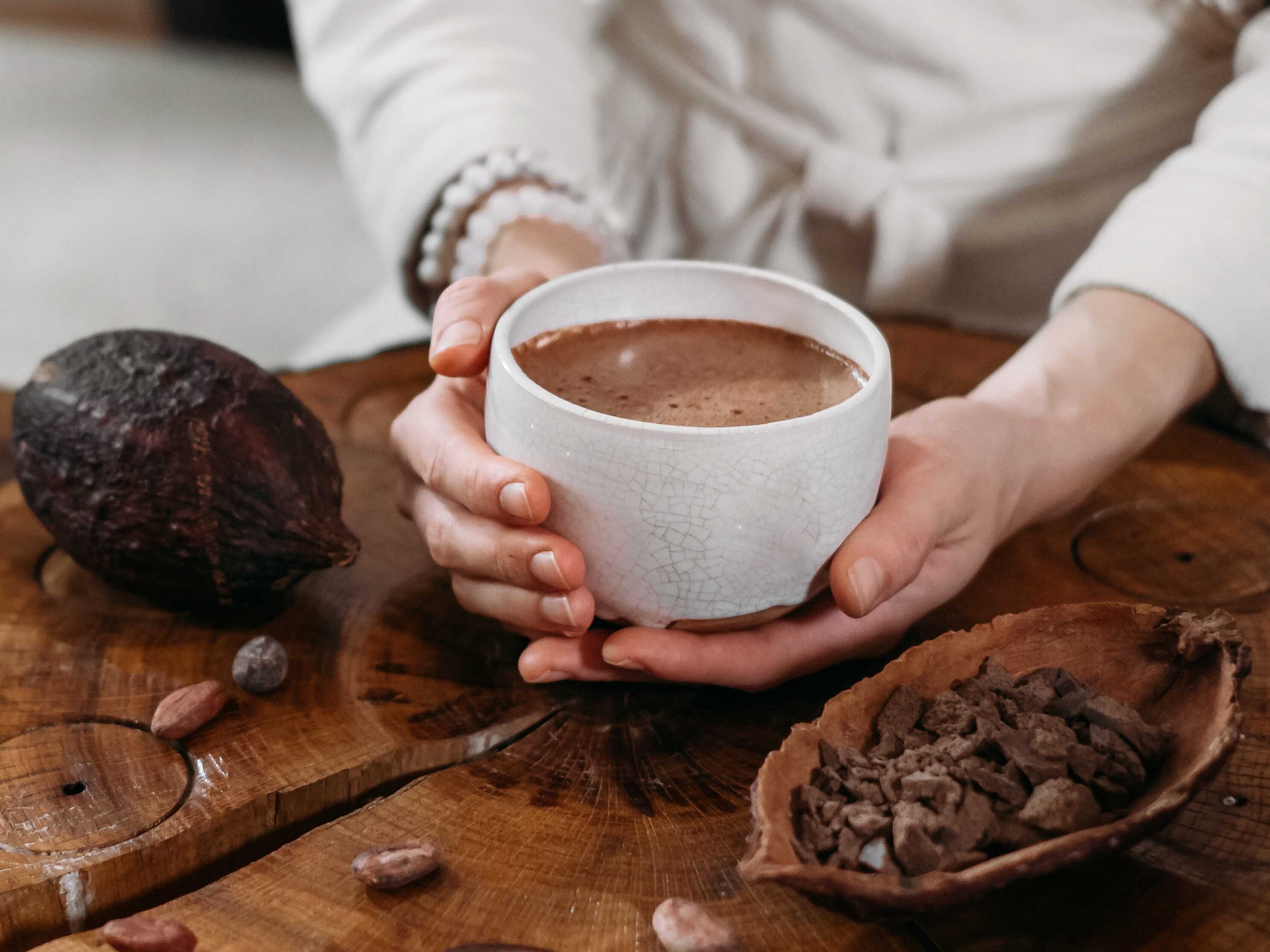 Eine Person hält eine Tasse mit Kakao in ihren Händen. Neben ihr liegen Kakeobohnen
